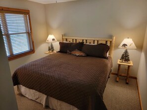 A bedroom with a bed, two bedside lamps, a window with blinds, and a carpeted floor.