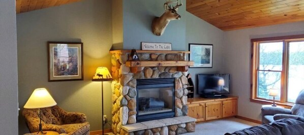 A cabin interior with a stone fireplace, a mounted deer head, a television, and a window with a view.