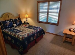 A bedroom with a bed, a nightstand, a lamp, a window with blinds, and a small wooden table.