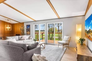 Ein modernes Wohnzimmer mit einer Couch, einem Sessel, einem großen Fenster mit Blick auf einen Garten und einem Bild an der Wand.