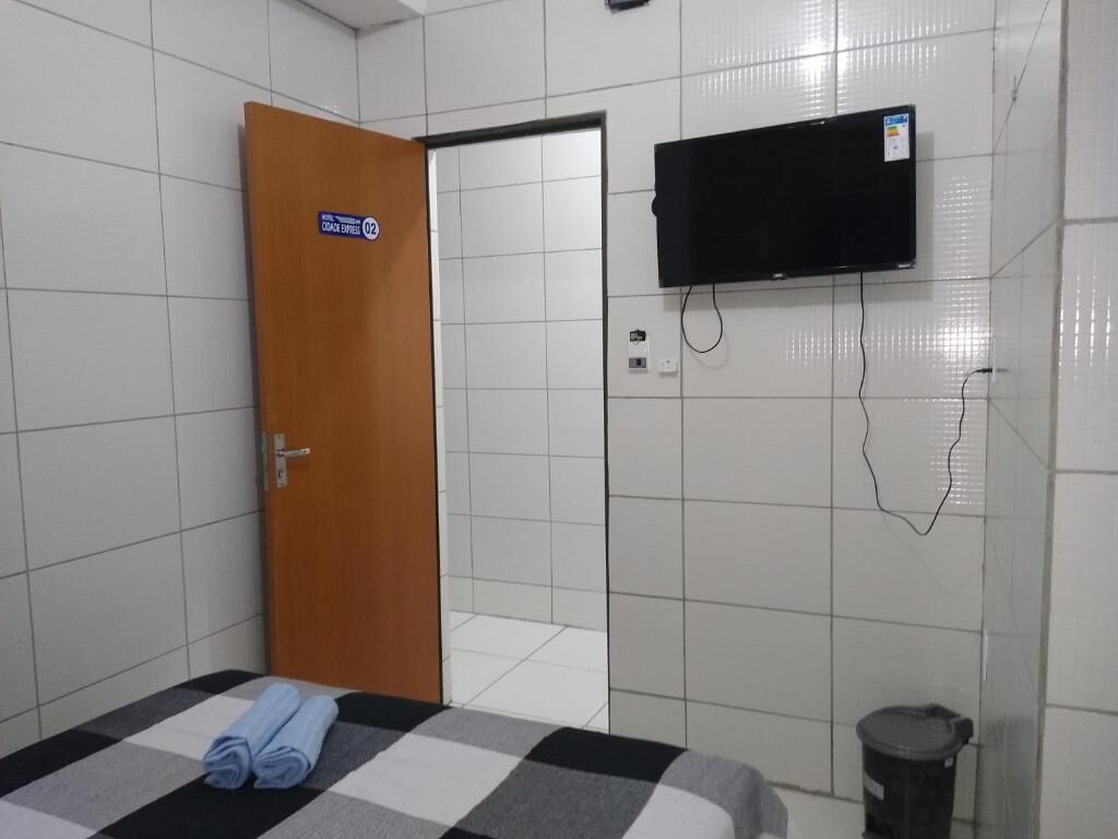 Una habitación de hotel con una mesa de recepción, una cama con colcha a cuadros, un televisor montado en la pared y un cubo de basura.