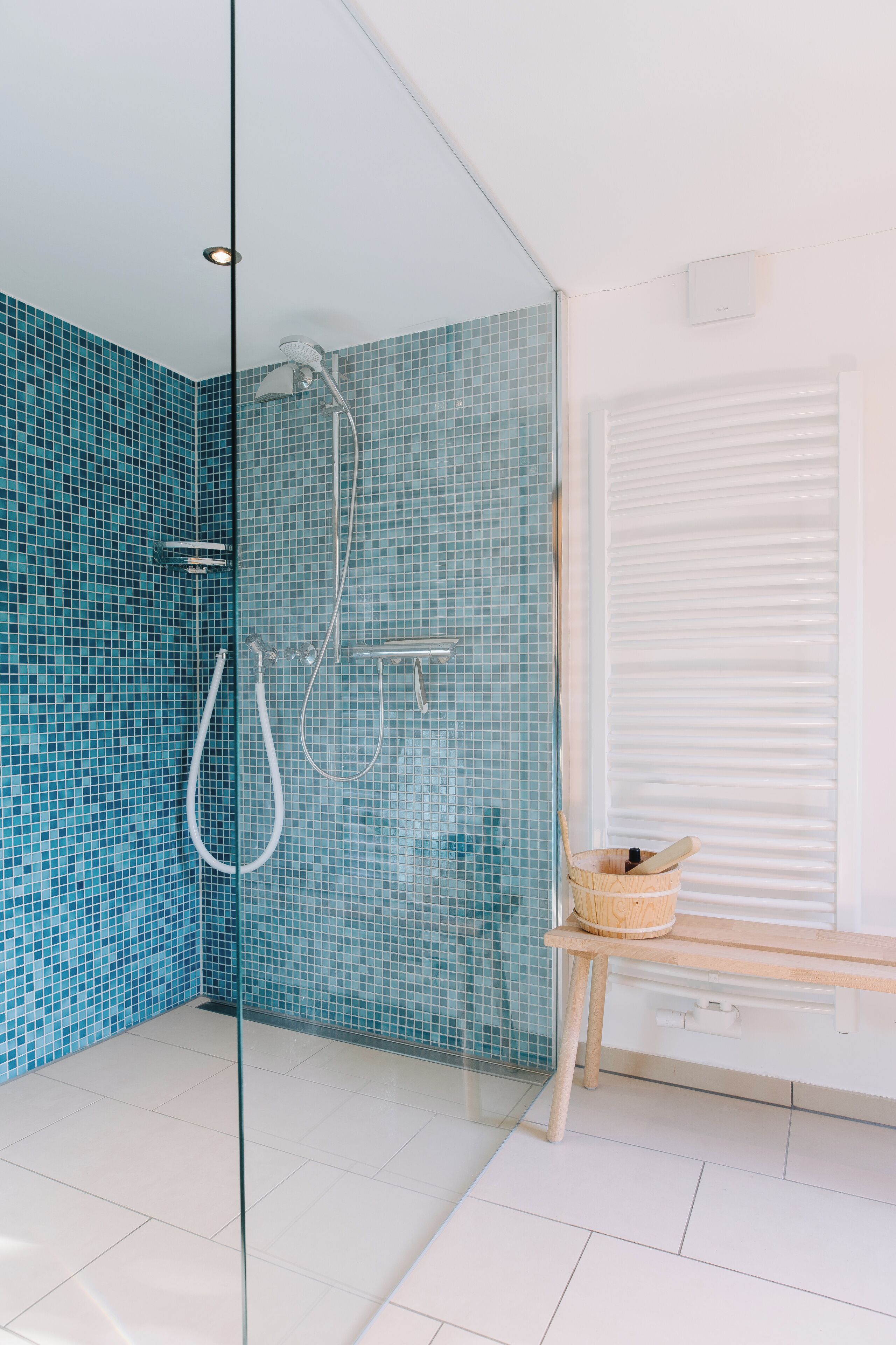 A bathroom with a glass partition, a shower with a blue mosaic tile wall, a wooden bench, and a white window with blinds.