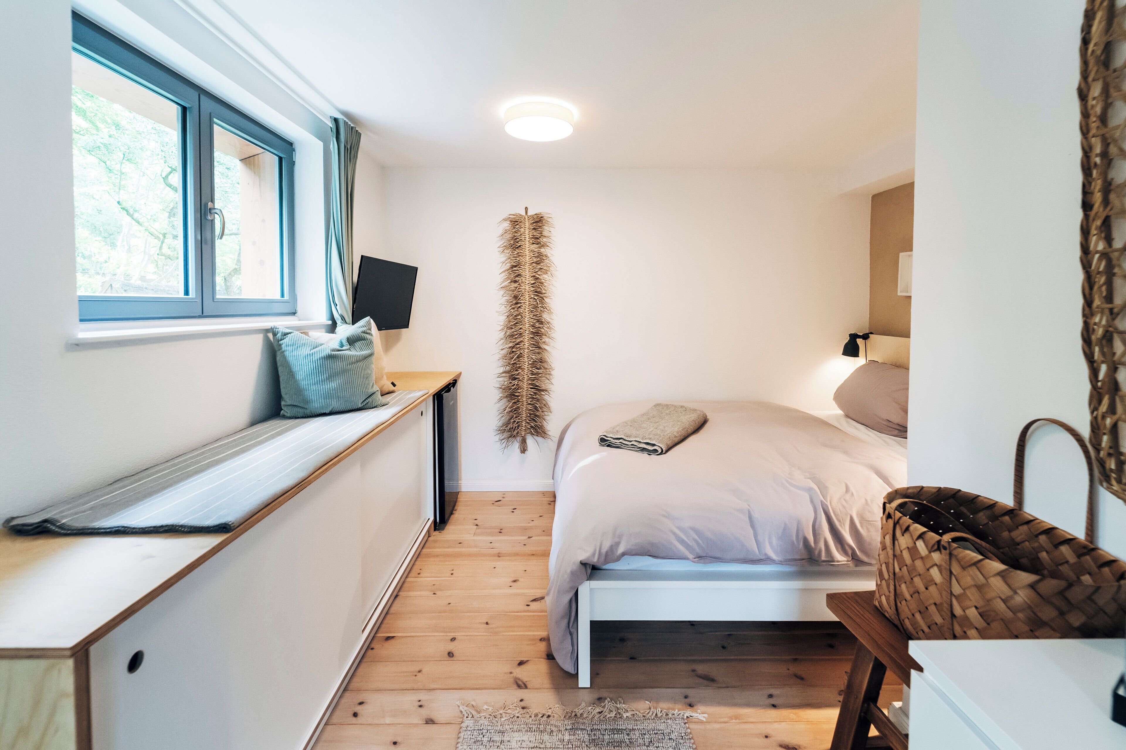 Un dormitorio con cama, ventana, escritorio, lámpara y cesta tejida.