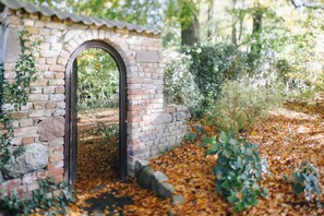 Ein steinerner Torbogen, der in einen Garten mit herabgefallenen Blättern und viel Grün führt.
