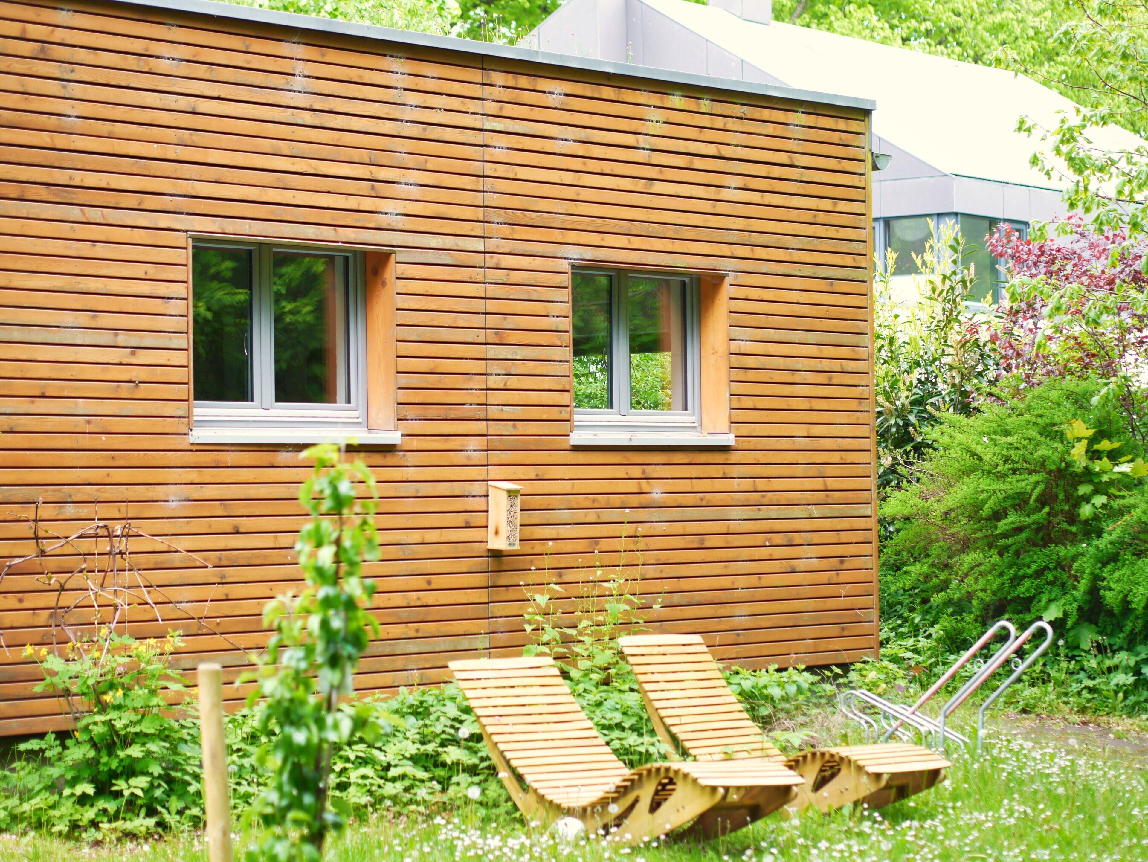 Eine Holzhütte mit zwei Fenstern, einer kleinen Veranda mit Holzliegen und einem Garten mit verschiedenen Pflanzen.