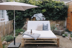 Ein Bett mit Kissen, einer Decke und einer Weinflasche auf einer Holzplattform, unter einem großen Sonnenschirm, umgeben von Grünpflanzen und einer Steinmauer.