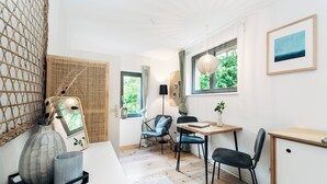 Ein Zimmer mit Holzboden, Schreibtisch, Stuhl, kleinem Tisch, einer Hängeleuchte, einem Fenster mit Blick auf Grünflächen und einem gerahmten Bild an der Wand.
