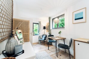 Ein Zimmer mit Holzboden, Schreibtisch, Stuhl, kleinem Tisch, einer Hängeleuchte, einem Fenster mit Blick auf Grünflächen und einem gerahmten Bild an der Wand.