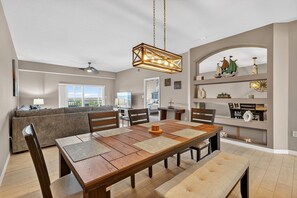 Ein Essbereich mit Holztisch, Stühlen und einer Bank. Es gibt einen Deckenventilator, eine Chandelierleuchtre und einen Blick ins Freie.
