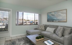 A living room with a large window, a grey sofa, a coffee table, and a painting on the wall.