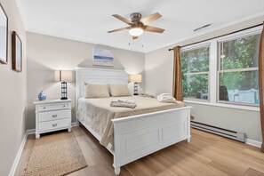 Ein Schlafzimmer mit Bett, Nachttischen, Deckenventilator, Fenster mit Vorhängen und Heizkörper.