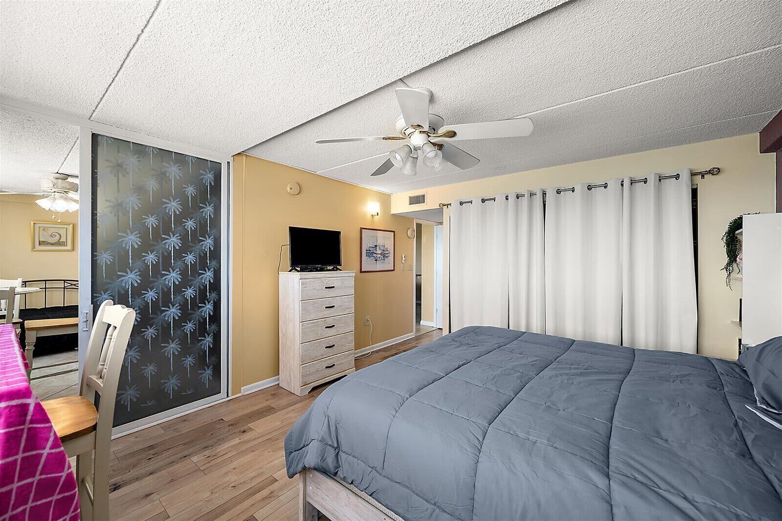 Un dormitorio con cama, cómoda, ventilador de techo y ventana con cortinas.