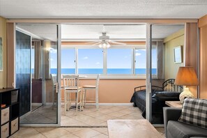 Ein Zimmer mit einem Bett, einem Sessel, einem Tisch, einer Lampe und Meerblick durch große Fenster.