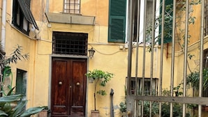 Un patio con un sendero de piedra que lleva a una puerta de madera, rodeado de edificios con paredes amarillas y contraventanas verdes.