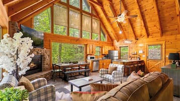 A log cabin interior with a stone fireplace, wooden dining area, kitchen, and large windows offering views of the forest.