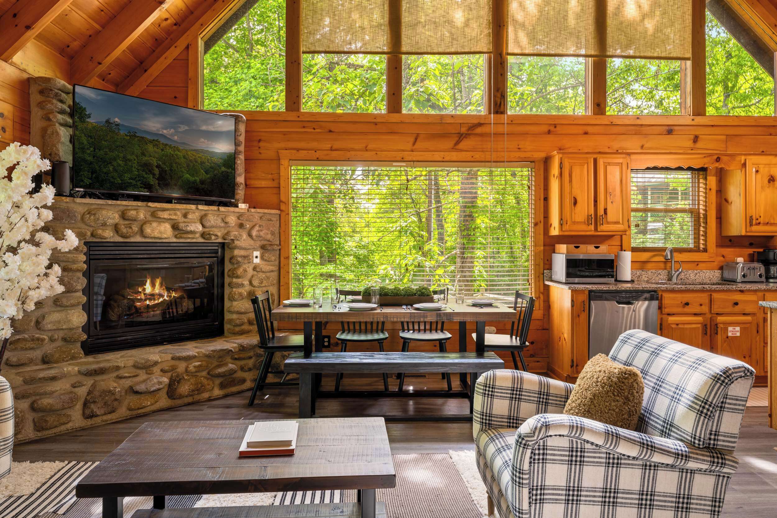 A cozy cabin interior with a stone fireplace, wooden dining table, and plaid sofa.