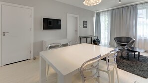 A modern living room with a white dining table, clear chairs, a flat-screen TV, a black leather chair, and a large window with curtains. - Valor by Grand Apartments (Sopot)