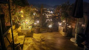 Vista nocturna de un balcón con plantas en macetas y luces de cuerda, con vistas a la ciudad.