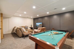 Ein Billardtisch mit Kugeln, eine Couch und ein Fernseher in einem Raum mit grauer Wand und Deckenleuchten.