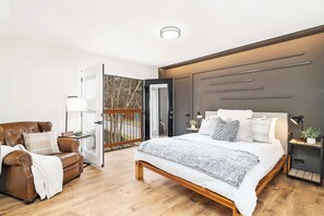 Una camera da letto moderna con un letto grande, una poltrona in pelle, un comodino con una lampada e una vista attraverso una porta aperta.