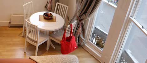 A cozy living room with a sofa, a dining table with chairs, a red bag, and large windows with curtains.