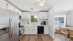 A modern kitchen with white cabinets, stainless steel appliances, a dishwasher, and a dining area with a wooden table and chairs. - Shore Retreat: 5 Bd home with alfresco living, hot tub, firepit, BBQ and games (Cape May)