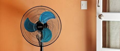Un ventilador de pie con base negra y aspas azules, un toallero blanco con perchas de madera y un mueble de madera con un interruptor de luz en una pared naranja.
