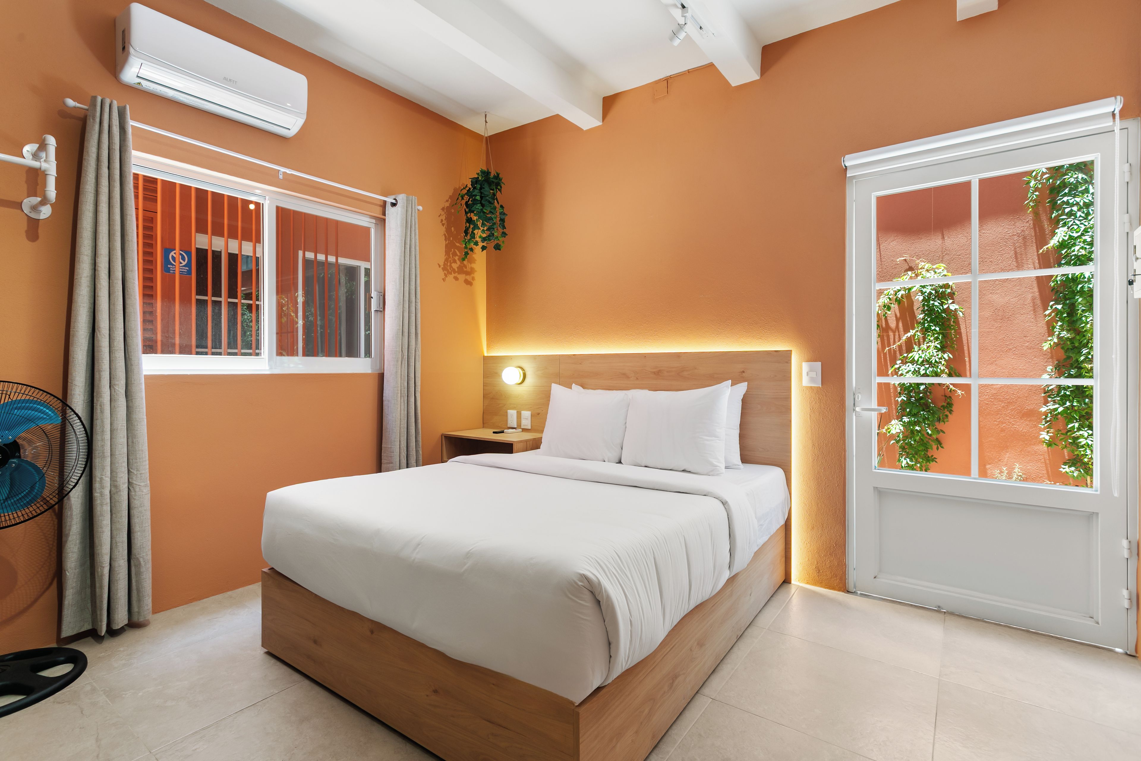 A hotel room with a bed, a bedside table with a lamp, a window with curtains, and an air conditioning unit.