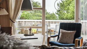 A cozy room with a large window, a blue armchair, a table with a candle, and a hanging light fixture.