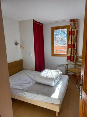 Ein Einzelbett mit Holz-Kopfstück, eine Wandleuchte, ein Fenster mit Blick auf ein Gebäude und ein Stuhl.