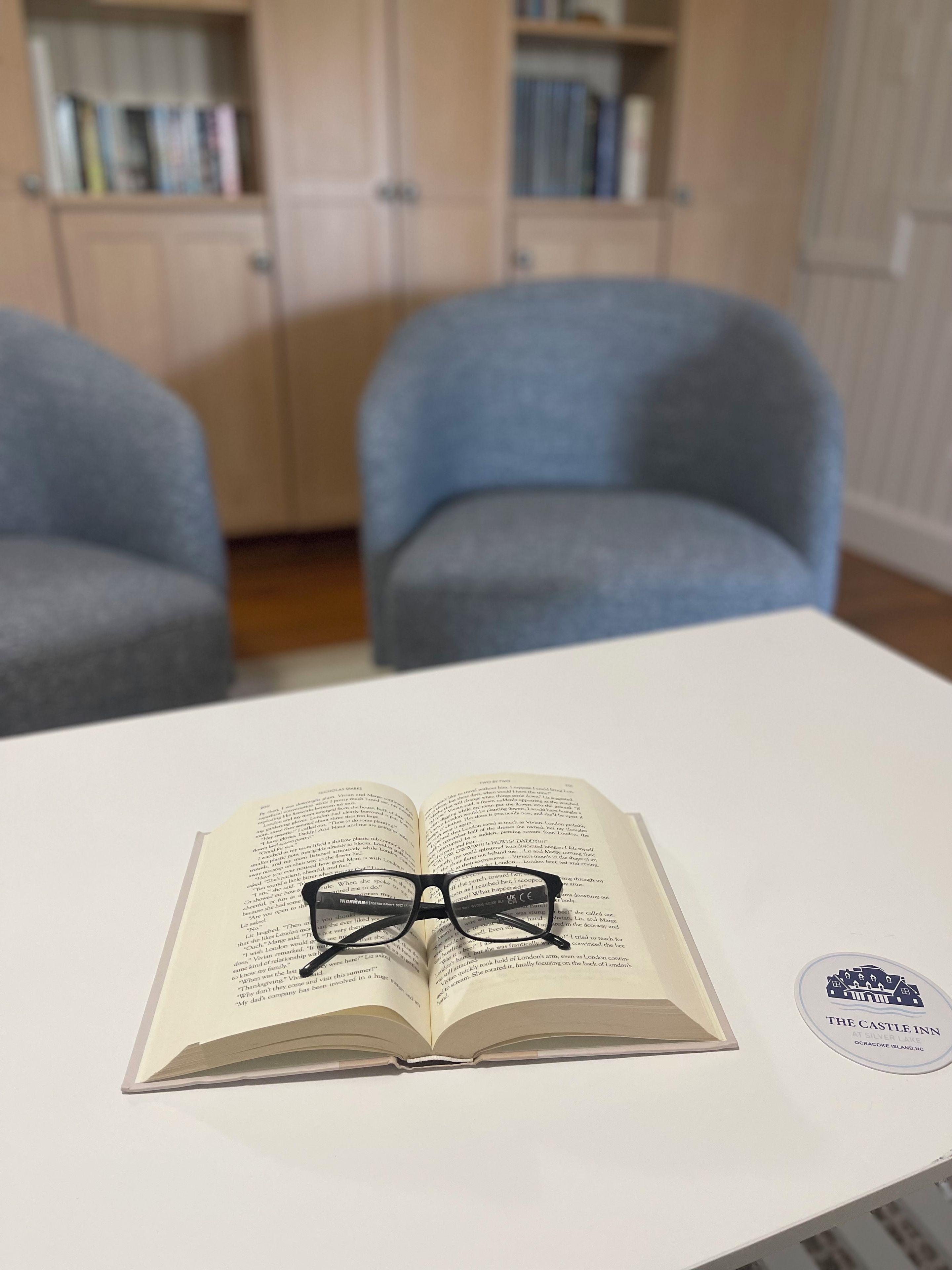 An open book with glasses on top, a white table, and a logo sticker on the table.