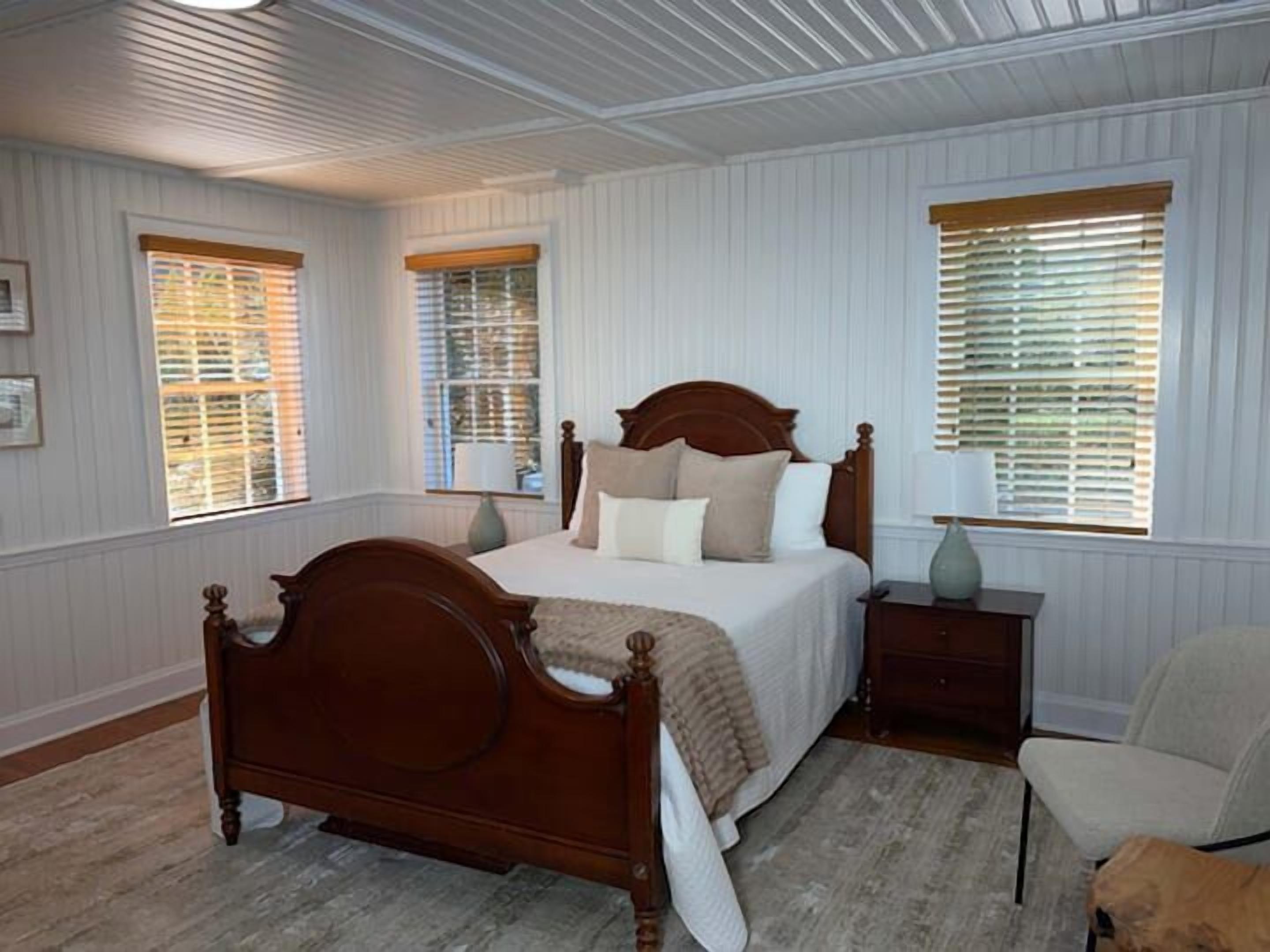A bedroom with a wooden bed, two bedside tables, a chair, and a window with blinds.