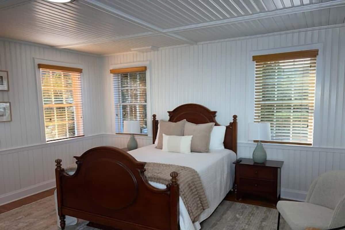 A bedroom with a wooden bed, two bedside tables, a chair, and a window with blinds.