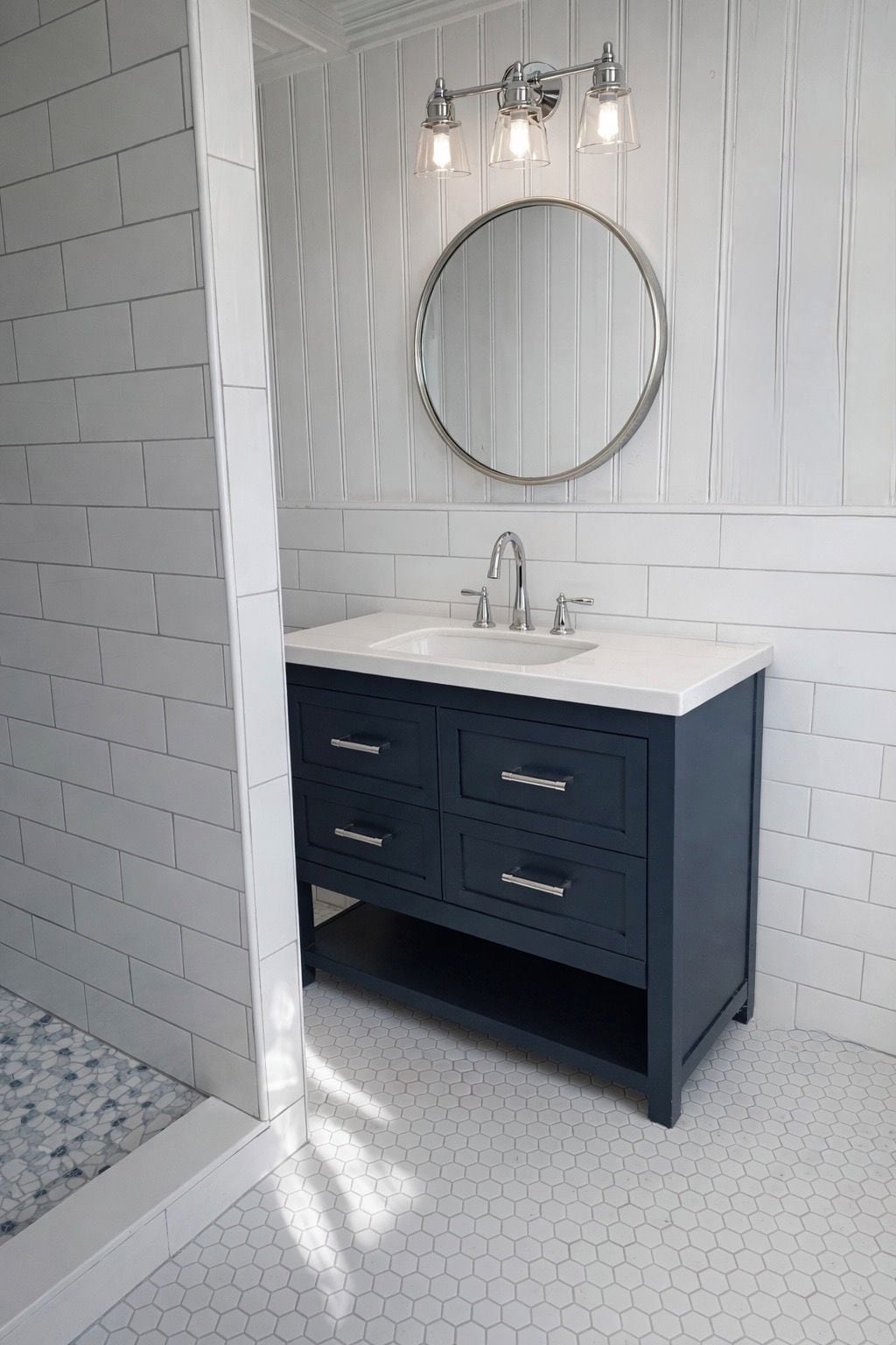 Une salle de bain avec un mur recouvert de carreaux blancs, un meuble-lavabo bleu foncé agrémenté d’un miroir rond et de trois lampes, et un sol carrelé avec des motifs hexagonaux.
