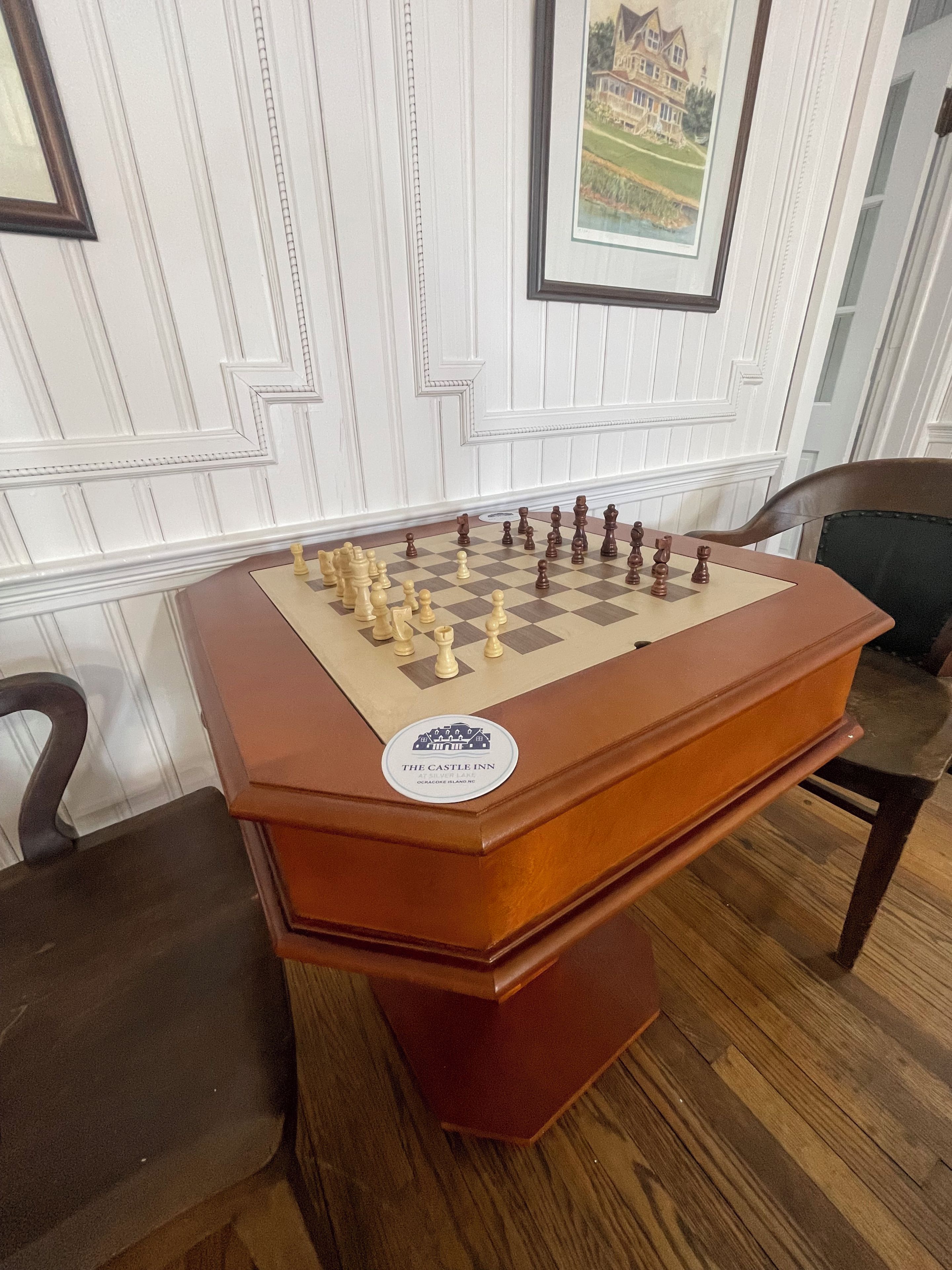 A chessboard on a wooden table with a logo sticker, set against a white paneled wall with a framed picture above.