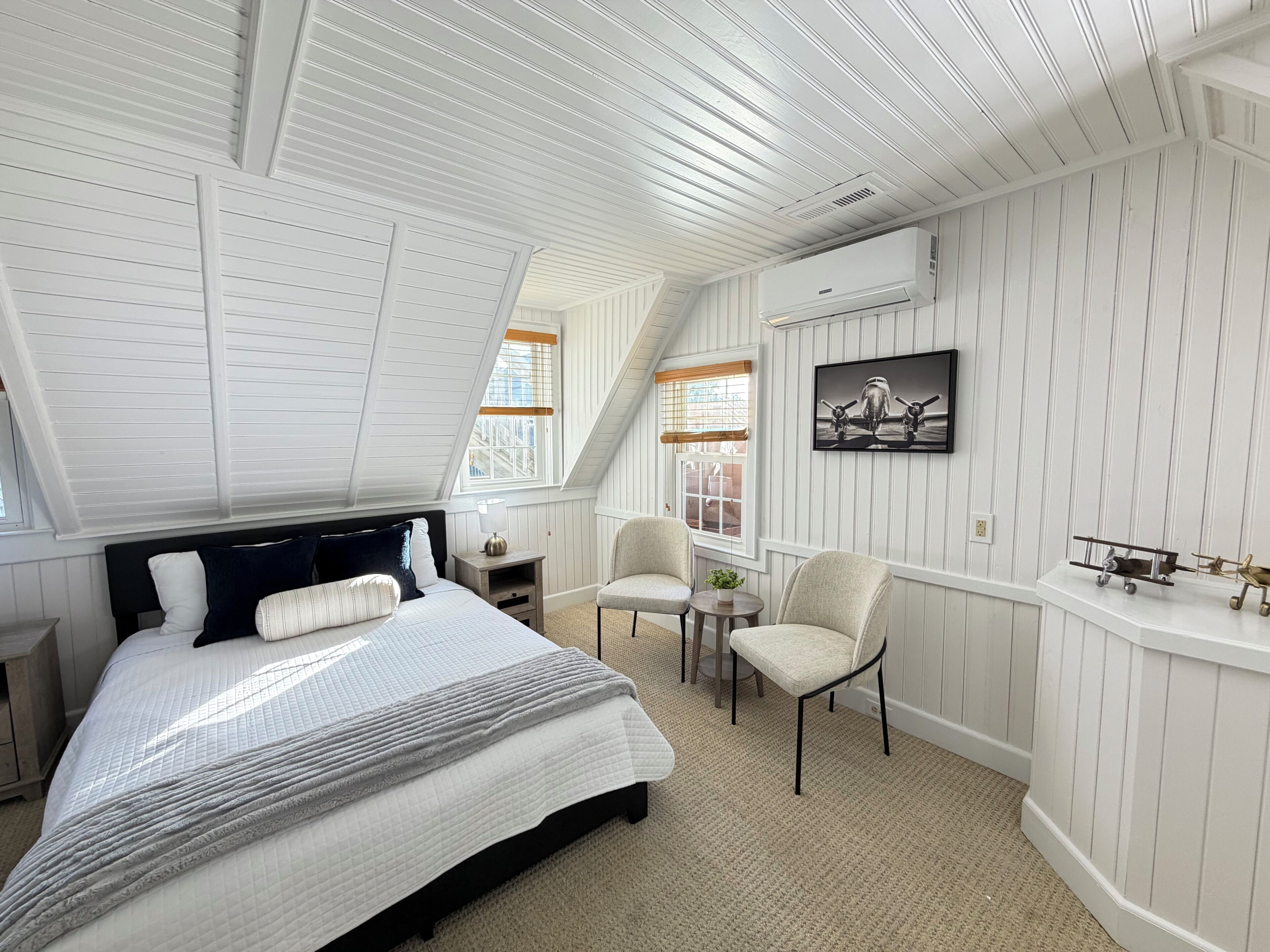 A bedroom with a sloped ceiling, a bed with a headboard, two chairs, a small table, and a wall-mounted air conditioner.