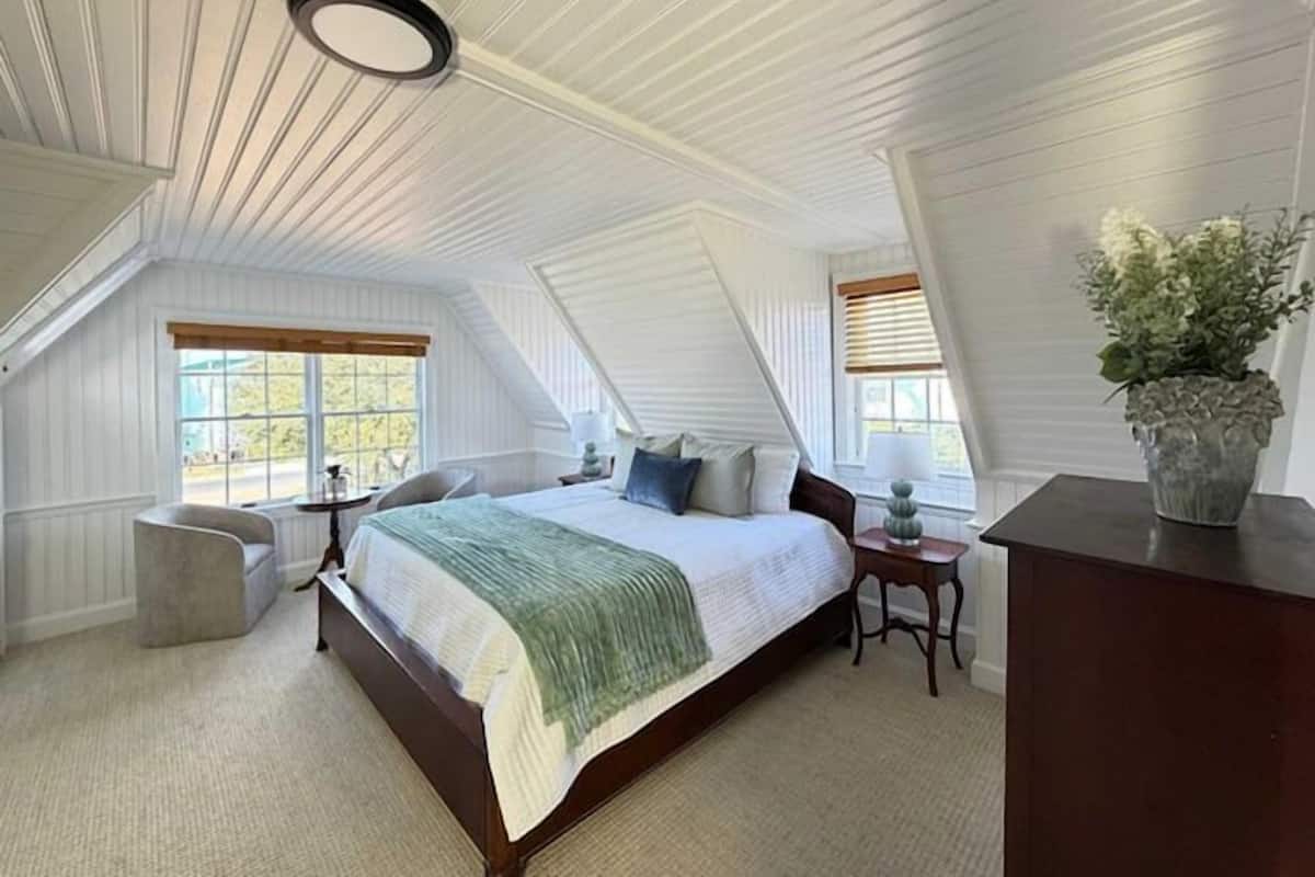 A bedroom with a sloped ceiling, a bed, a nightstand, a chair, a desk, and a potted plant.