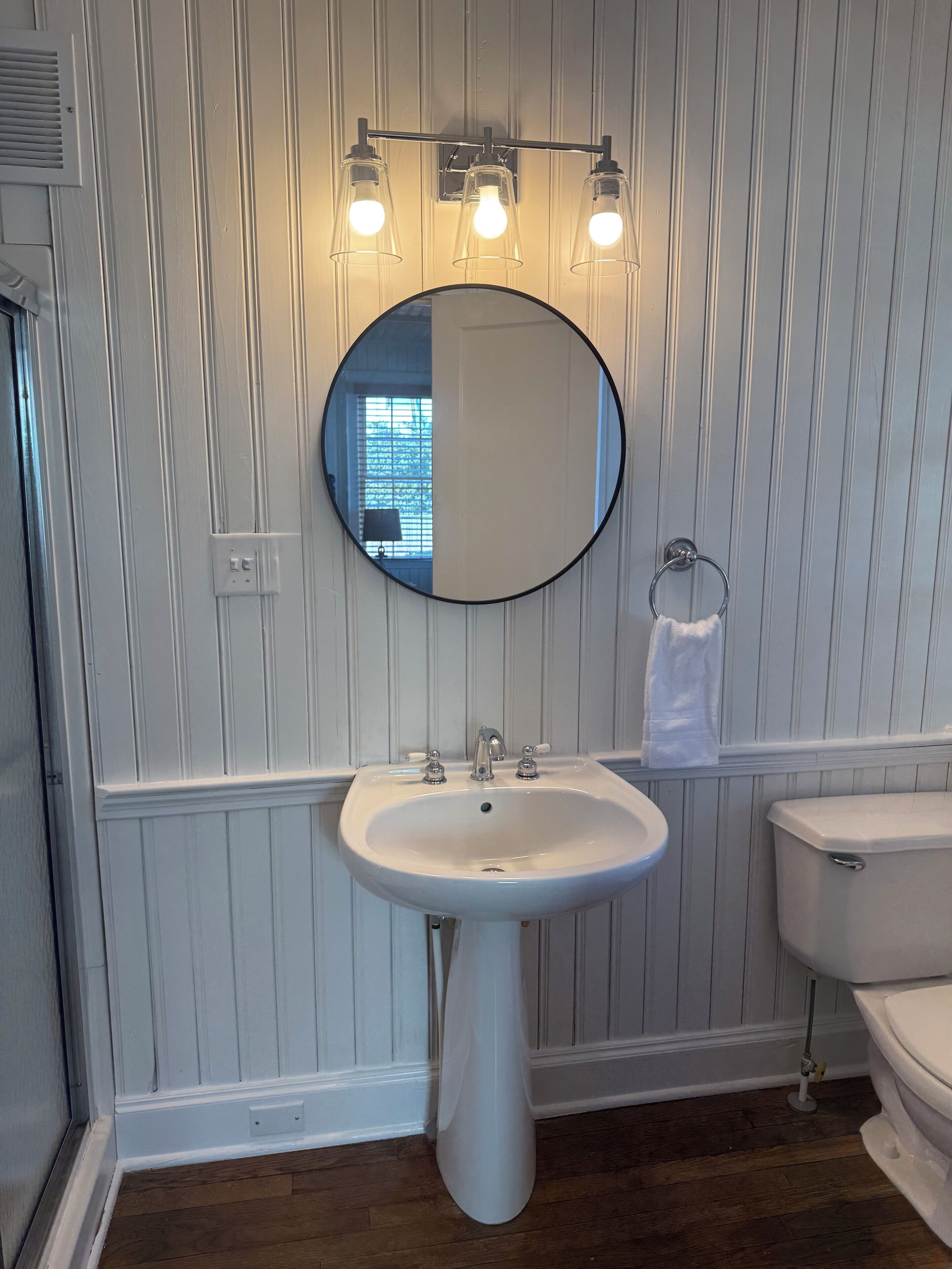 Une salle de bain avec un lavabo blanc, un miroir rond et des toilettes. Trois luminaires sont fixés au-dessus du miroir.