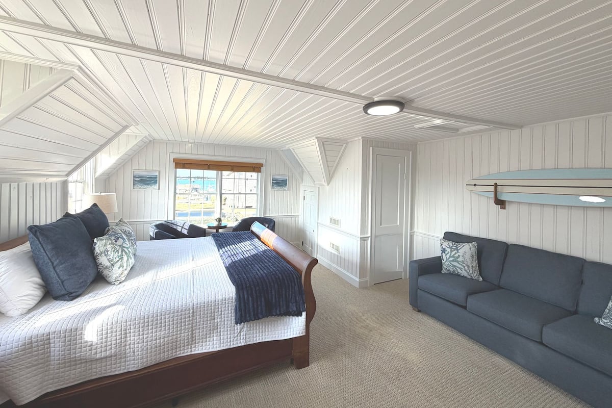 A bedroom with a bed, a sofa, a window, and a surfboard on the wall.