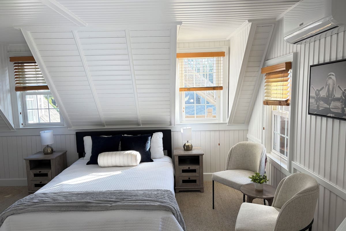 A bedroom with a sloped ceiling, a large bed, bedside tables, a chair, and a window with blinds.
