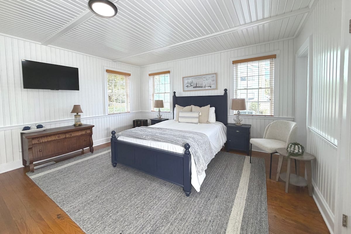 A bedroom with a bed, nightstands, a dresser, a TV, and windows with blinds.