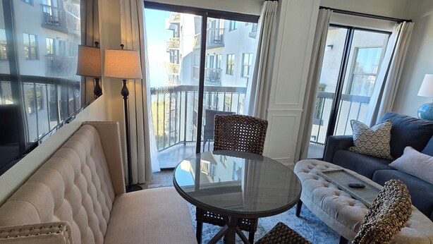 A hotel room with a sofa, a round table, wicker chairs, a glass-top table, and a balcony with a view of buildings. - Lakeside Universal Retreat (Orlando)