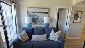 A living room with a dark blue sofa, a round ottoman, a remote control on the ottoman, and a painting of a beach scene on the wall. - Lakeside Universal Retreat (Orlando)