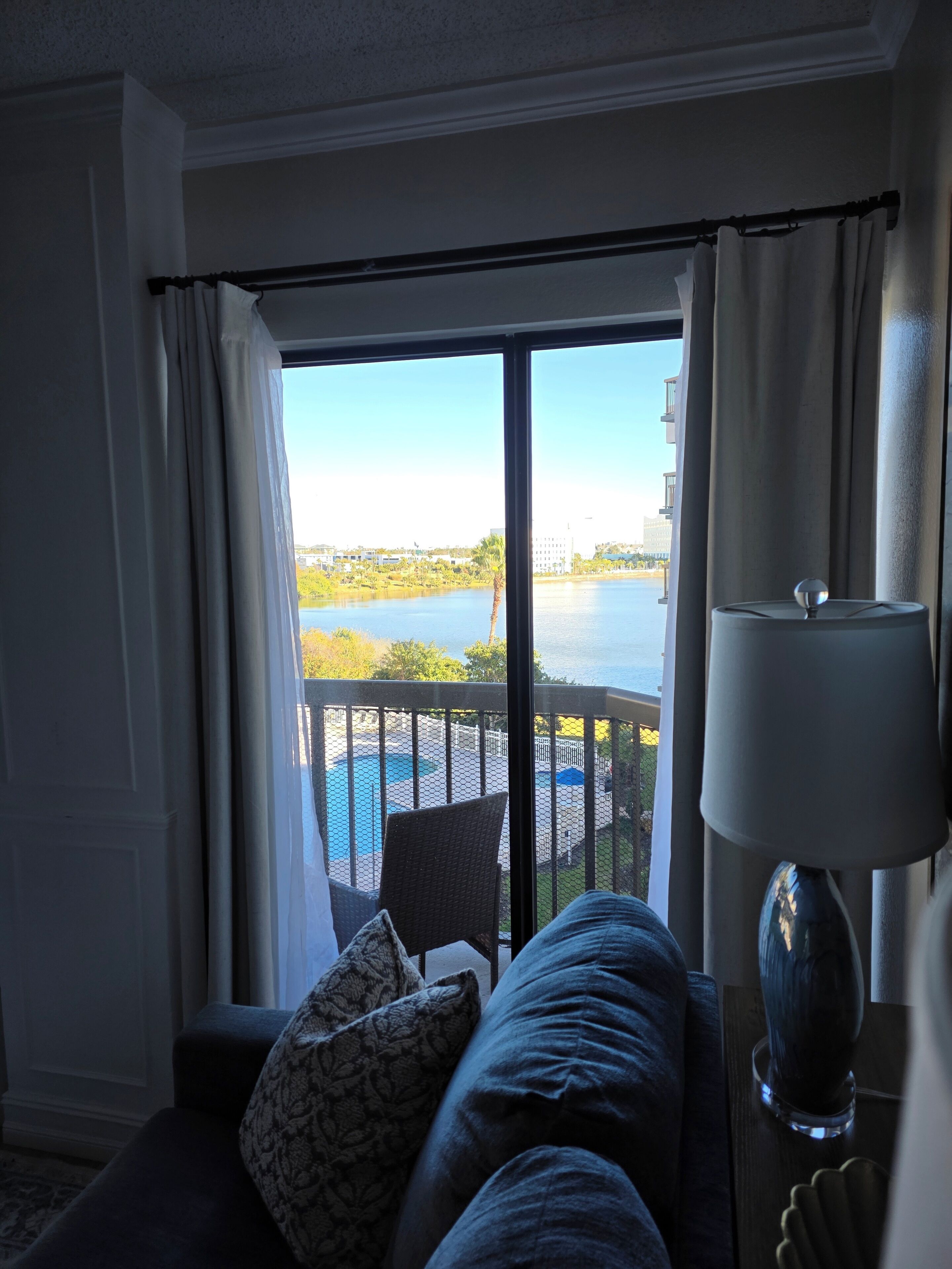 A room with a view of a swimming pool and a body of water, a sofa with cushions, and a lamp.