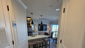 A modern kitchen with a central island, a dining area with bar stools, a flat-screen TV mounted on the wall, and a view of the outdoors through a window. - Lakeside Universal Retreat (Orlando)