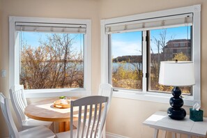 Ein Zimmer mit zwei Fenstern, einem Tisch mit Stühlen, einer Lampe und Blick auf Bäume und Gebäude.