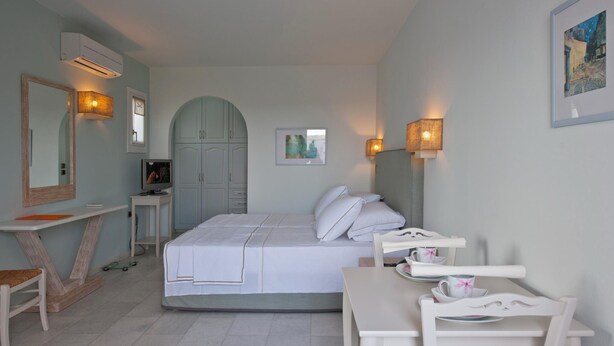 A bedroom with a bed, bedside table, chair, desk, and an arched doorway. - Double Suite 1 balcony (Naxos)
