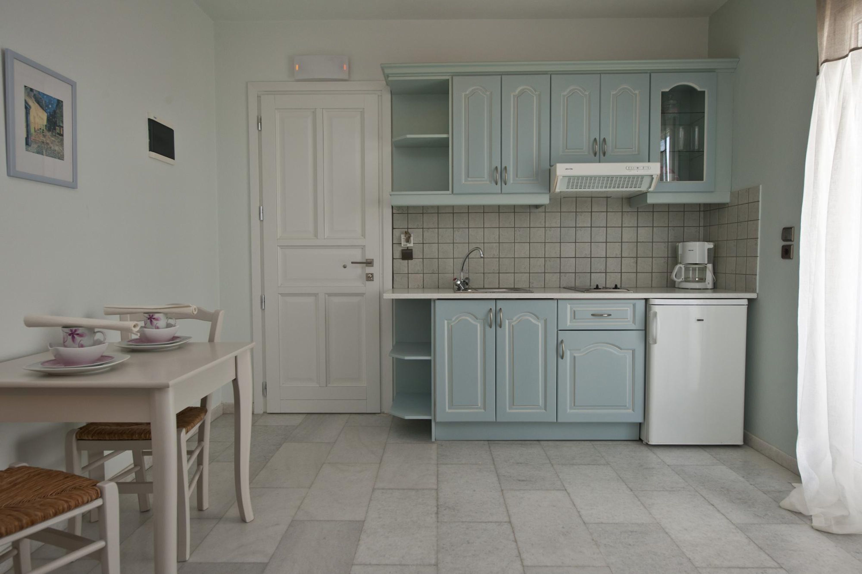 Eine Küche mit hellblauen Schränken, einem weißen Kühlschrank, einer Kaffeemaschine und einem Esstisch mit Stühlen.
