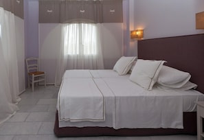 Ein Doppelbett mit weißen Leinen, ein Sessel, eine Wandlampe und ein Fenster mit durchscheinenden Vorhängen.