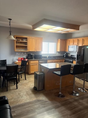 A modern kitchen with wooden cabinets, a central island, bar stools, and a dining area with a table and chairs.